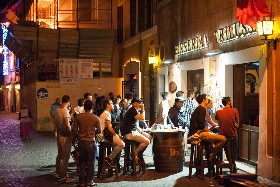 Personnes autour de tables-tonneaux devant la Birreria Trilussa le soir, dans le Trastevere à Rome